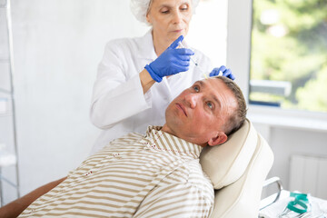 Obraz premium Adult man undergoing botulinum therapy session at aesthetic medicine clinic. Skilled woman cosmetologist making injections between eyebrows of patient with insulin syringe to correct facial wrinkles