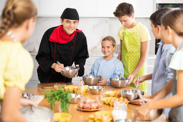Enthusiastic young chef giving culinary classes to group of interested curious tween girls and boys, teaching to cook deliciously..