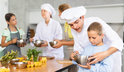 Focused interested preteen girl mixing sauce in metal bowl with whisk under guidance of friendly professional young chef during cooking masterclass for children