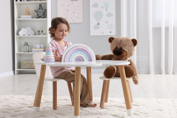 Cute little girl playing with toy and teddy bear at white table in room