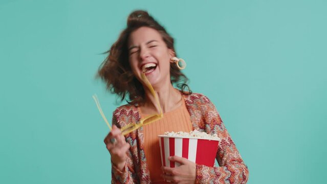 Excited young woman in 3D glasses eating popcorn and watching interesting tv serial, sport game, film, online social media movie content. Brunette pretty girl isolated on blue studio background