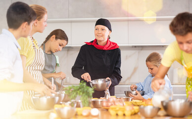 During lesson at cooking course, woman cook in black suit and hat tells children about rules of cooking fluffy pancakes. Female professional shows example of successful mobile dough