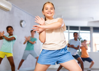 Active preteen girl practicing sport dances with other children in training hall during dancing...