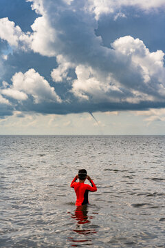 A boy wading in the ocean watches as a funnel cloud descends to the horizon in the distance