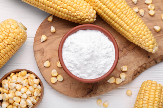 Bowl with corn starch, ripe cobs and kernels on white wooden table, flat lay