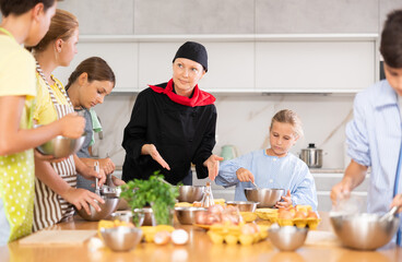 During lesson at cooking course, woman cook in black suit and hat tells children about rules of cooking fluffy pancakes. Female professional shows example of successful mobile dough