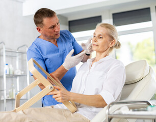 Obraz premium Interested senior female patient sitting on couch in medical aesthetic office looking at mirror while professional male cosmetologist explaining future hardware rejuvenating facial procedure