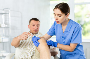 Obraz premium Patient man is sitting on medical couch, stroking sore knee with hand and telling young female doctor complaints about how he feels