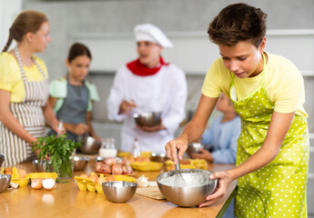 During culinary classes, teen boy combine essential components in mixing bowl and readies dough for bread rolls. In background, blurry children stand near kitchen table and listen to chef explanations