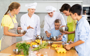 During lesson in cooking courses, female cook professional tells children about rules ticks and tricks for making fluffy pancakes, guy assistant helps hesitant child mix ingredients in bowl