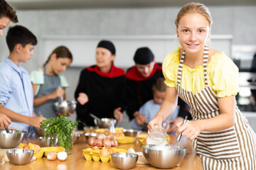 During cooking classes, girl carefully mixes ingredients in bowl with whisk. In background, blurry children stand near kitchen table and listen to chefs explanations