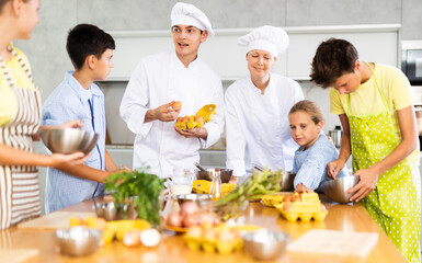 Amiable woman and young man, skilled chefs wearing white cook jackets and toques, running culinary courses for preteen children, sharing secrets of cooking