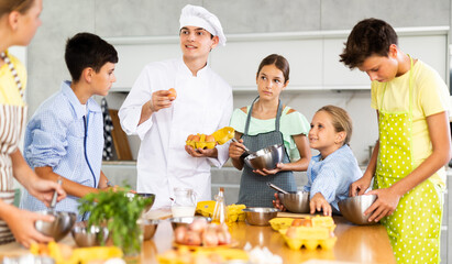 Young guy cook in uniform teaches group of children how to cook dish