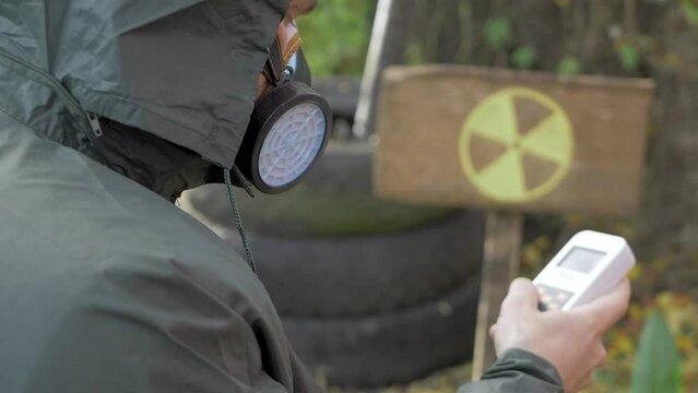 Use radiation geiger counter, checks level of radiation. Yellow danger sign in radioactive zone background. Nuclear weapons, atomic apocalypse concept. Male in protective suit and mask respirator 