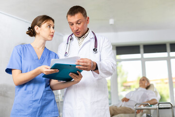 Two professional doctors, adult man and young woman, standing in office, reading medical card of patient and discussing diagnosis..