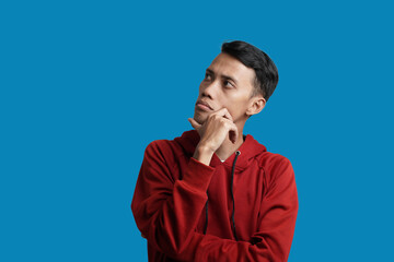 Portrait of young Asian man wearing red hoodie thinking and looking for solution, isolated on blue background.