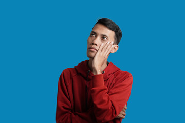 Portrait of young Asian man wearing red hoodie thinking and looking for solution, isolated on blue background.