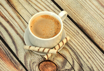 A cup of fresh cappuccino with foam and two waffle rolls on a saucer on a light wooden table.