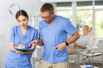 Two fellow doctors, woman and man, standing in medical office with papers in hands, focused on...