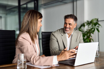 Happy older Indian business man professional board executive and mature female manager wearing suits discussing corporate management data using laptop sitting at table in office at meeting.