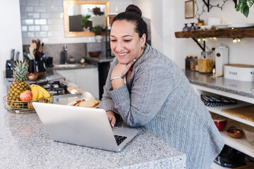 young latin woman overweight using laptop while having breakfast at home kitchen in Mexico Latin...