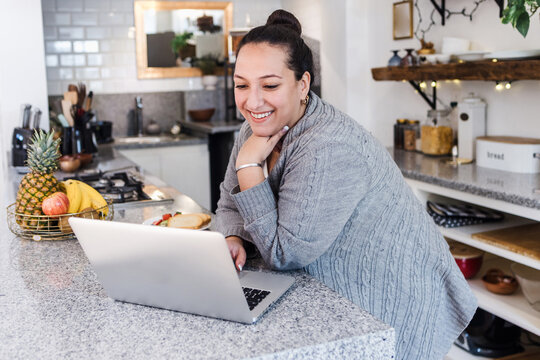 young latin woman overweight using laptop while having breakfast at home kitchen in Mexico Latin America, hispanic plus size female