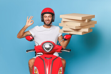 Bearded man in red helmet sits on red scooter in front of blue background, one hand shows ok, in other hand boxes with pizza, delivery concept, copy space © South House Studio