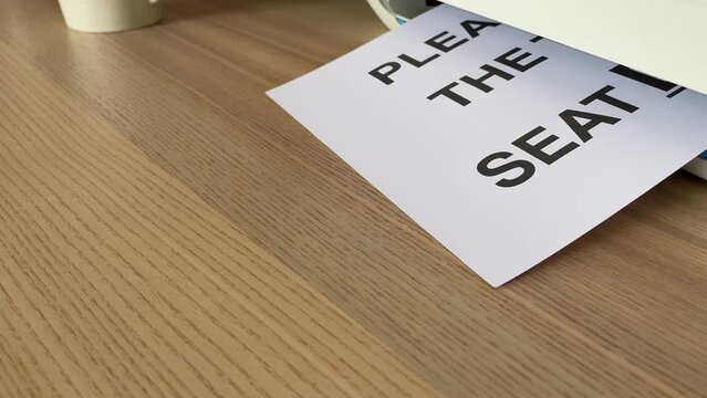 &ldquo;Please put the toilet seat down&rdquo; black text word on paper sheet A4 document sliding out of printer scanner on wooden table close up view