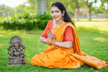 Happy young indian woman wearing saree sitting worshiping lord Ganesha idol outdoor at park...