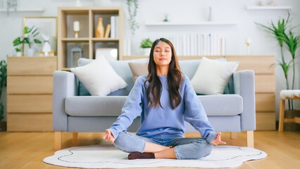 Happy young Asian woman practicing yoga and meditation at home sitting on floor in living room in lotus position and relaxing with closed eyes. Mindful meditation and wellbeing concept - obrazy, fototapety, plakaty