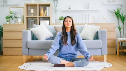 Fototapeten Happy young Asian woman practicing yoga and meditation at home sitting on floor in living room in lotus position and relaxing with closed eyes. Mindful meditation and wellbeing concept © Monster Ztudio
