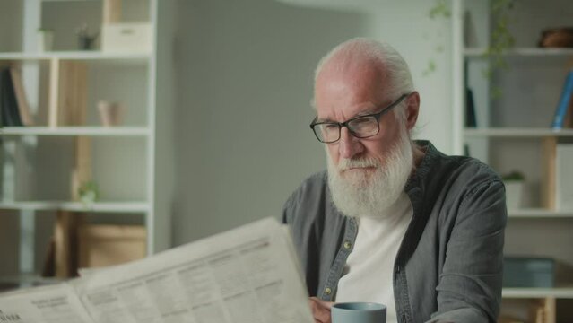 A Serious, Smart Old Man Sits at a Table and Reads a Newspaper.An Elderly Man with a Newspaper, Drinking Coffee Alone at Home, Sees the News and Current Events in the Newspaper.Daily Newspaper Concept
