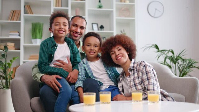 Portrait of smiling african american adults and kids in casual clothes sitting on couch in spacious room. Handsome dad holding son on knees while mom snuggling up with younger boy for family picture