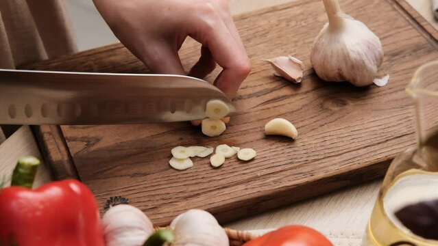 Close-up of a woman cutting fresh garlic with a knife in the kitchen. Preparing vegetable salad. Housewife chopping garlic with knife on wooden cutting board