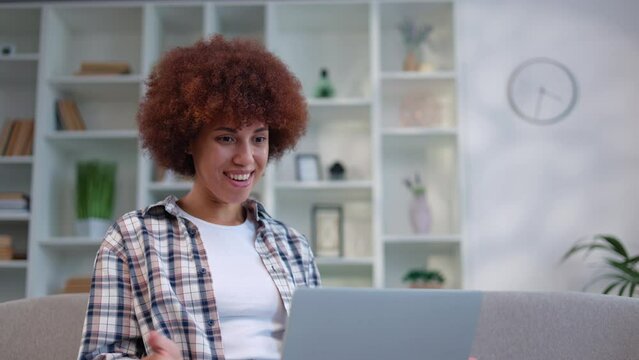 Positive multiracial woman with curly hairstyle waving hand and talking during video conference on laptop. Young female sitting on grey couch at living room and conduction work meeting online