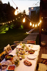 Row of lamps hanging over long table served with appetizing homemade food and drinks, fresh fruits,...