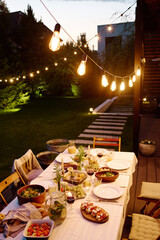 Row of lamps hanging over long table served with appetizing homemade food and drinks, fresh fruits, plates, burning candles and flowers in vases