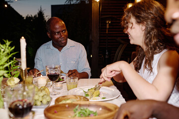 Young brunette woman sitting next to mature African American man by table served with homemade food, eating and listening to him by dinner
