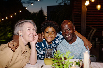 Mature intercultural couple and their cute grandson looking at camera while sitting by served table in the evening and enjoying weekend
