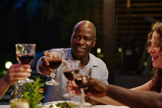 Focus on mature African American man with glass of red wine toasting with guests and clinking with their wineglasses during family dinner