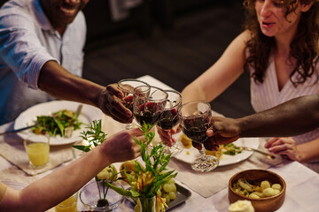 Close-up of several intercultural familiy members clinking with glasses of homemade red wine while sitting by table served with appetizing food