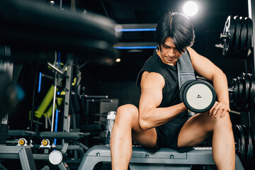 Fototapeta na wymiar A handsome young Asian man sitting on an exercise bench holding a heavy dumbbell, showcasing his muscular arm and bicep, and his dedication to fitness. empty space