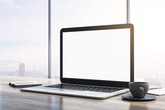 Close up of laptop computer with empty white mock up monitor, coffee cup and notepad placed on wooden desktop. Window with bright city view in the background. 3D Rendering.