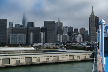 Fototapeta na wymiar Grey foggy low hanging clouds day in San Francisco Bay with port piers, city skyline downtown architecture highrise buildings and landmarks as Coit Tower or Pyramid