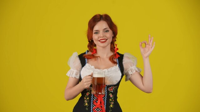 Medium yellow background isolated shot of a young German woman, waitress, wearing a traditional costume, holding a glass of lager, beer, showing OK sign and smiling.