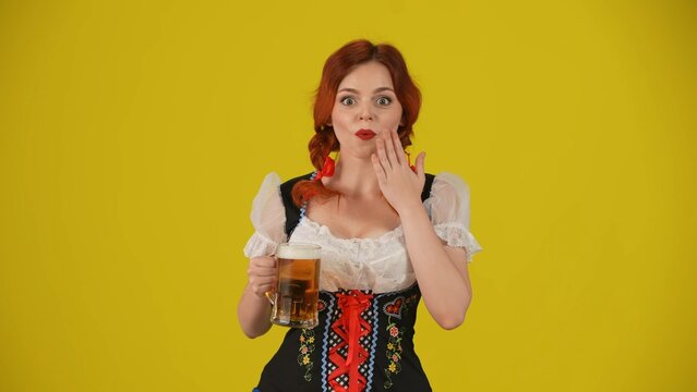 Medium yellow background isolated shot of a young German woman, waitress, wearing a traditional costume, holding a glass of lager, beer, expressing wow and excitement.