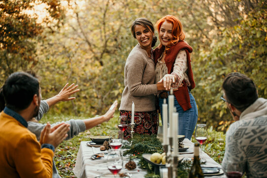 The joyous ambiance of a garden party, with a lesbian proposal with a diverse group of people relishing a shared meal amidst nature's beauty.