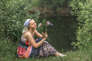 A beautiful young woman with blond hair in a sundress on the shore of a lake with a bouquet of wildflowers. National Russian clothes . Country life