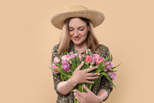 Happy young woman in straw hat holding bouquet of beautiful tulips on beige background