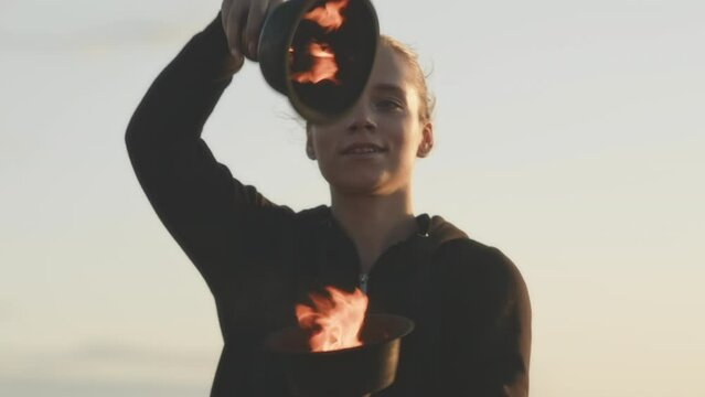 Medium shot of artistic Caucasian girl dancing with fire in bowls at sunset, making smooth movements with her hands